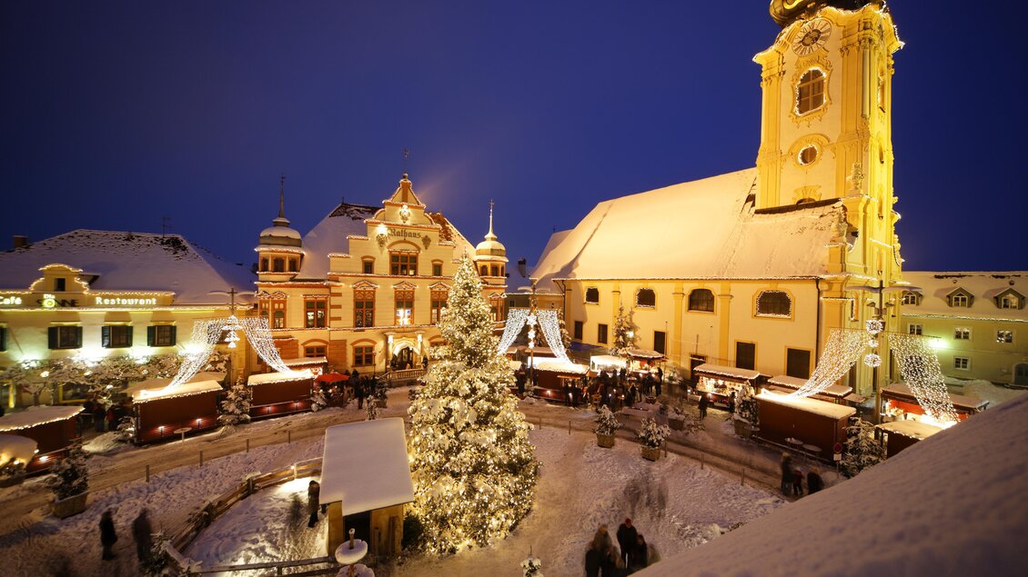 Ein festlicher Weihnachtsmarkt mit festlich beleuchteten Ständen und einem großen, geschmückten Baum. Der Platz ist von schneebedeckten Gebäuden und einer Kirche umgeben, alles unter einem klaren Nachthimmel. | © Bernhard Bergmann