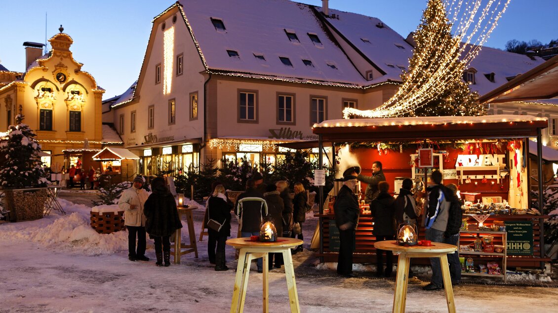 Eine festliche Weihnachtsmarkt-Szene mit geschmückten Ständen und vielen Menschen. Im Hintergrund sind mit Schnee bedeckte Häuser und Lichter zu sehen. | © Bernhard Bergmann