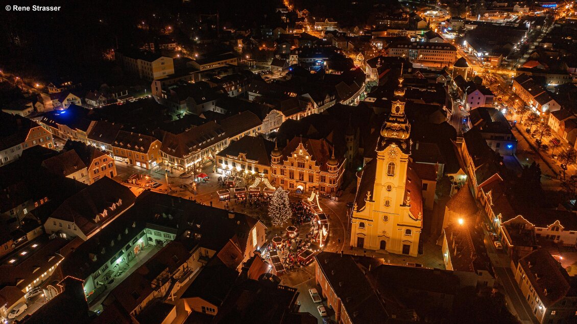 Eine festliche Stadtansicht bei Nacht mit beleuchteten Gebäuden und einem Weihnachtsmarkt. Im Mittelpunkt steht ein großer Weihnachtsbaum und die Straßen sind festlich dekoriert. | © Rene Strasser