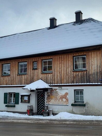 The multi-storey, traditional wooden house of the tavern "Postillions Einkehr" in Altaussee with a snow-covered roof. In front of the inn lies a small paved area with a white fence. | © Petra Kirchschlager