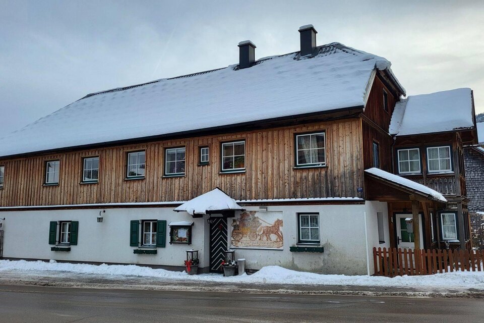 Das mehrstöckige, traditionelle Holzhaus der Postillions Einkehr in Altaussee mit einem schneebedeckten Dach. Vor dem Gasthaus liegt eine kleine, gepflasterte Fläche mit einem weißen Zaun. | © Petra Kirchschlager