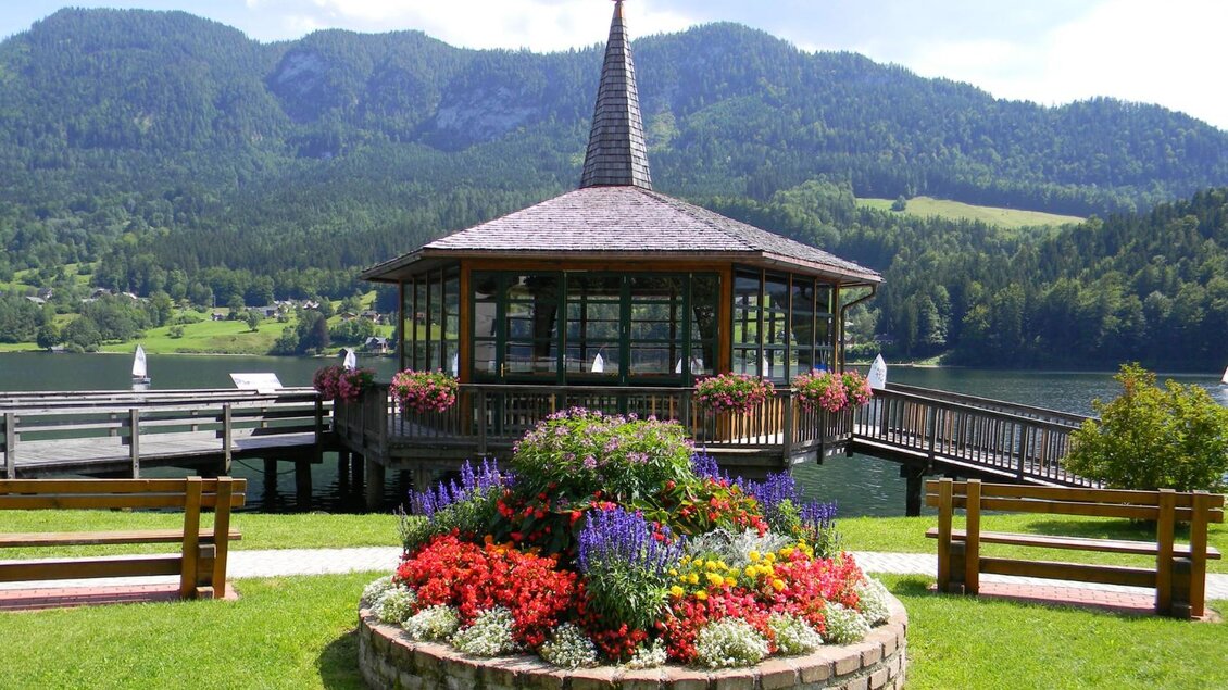 Eine malerische Seeterrasse mit einem Pavillon und bunten Blumenbeeten. Im Hintergrund erheben sich grüne Berge unter einem klaren Himmel. | © Gemeinde Grundlsee