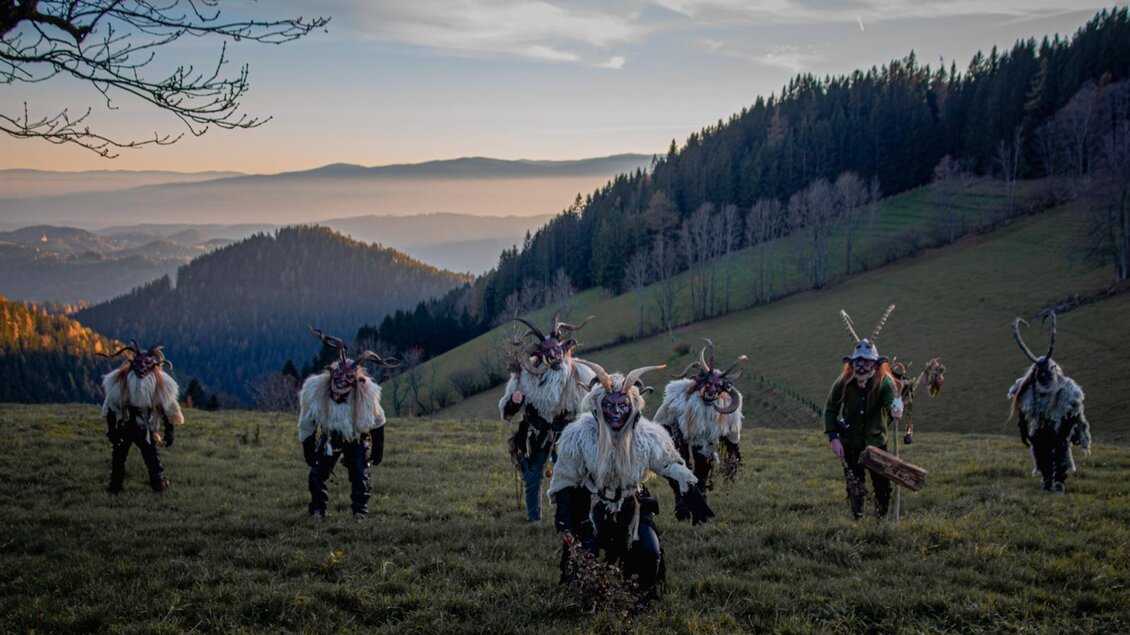 Eine Gruppe von Personen in traditionellen Kostümen mit Masken läuft über eine Wiese in den Bergen. Der Hintergrund zeigt Hügel und einen klaren Himmel in der Dämmerung. | © Übelbacher Gleinalm Pass
