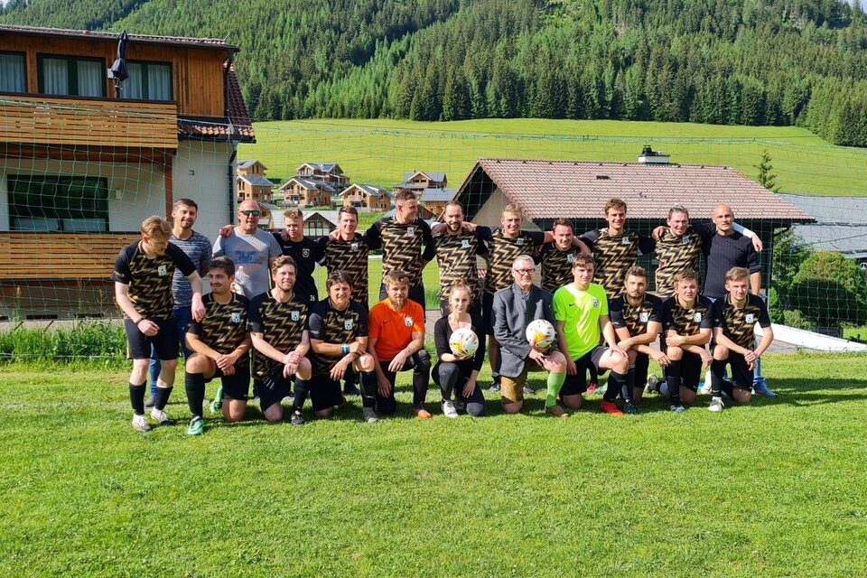 Eine Fußballmannschaft steht auf einem Rasenplatz vor einer malerischen Berglandschaft. Im Hintergrund sind Hütten und Bäume zu sehen. | © STR Brodjäger