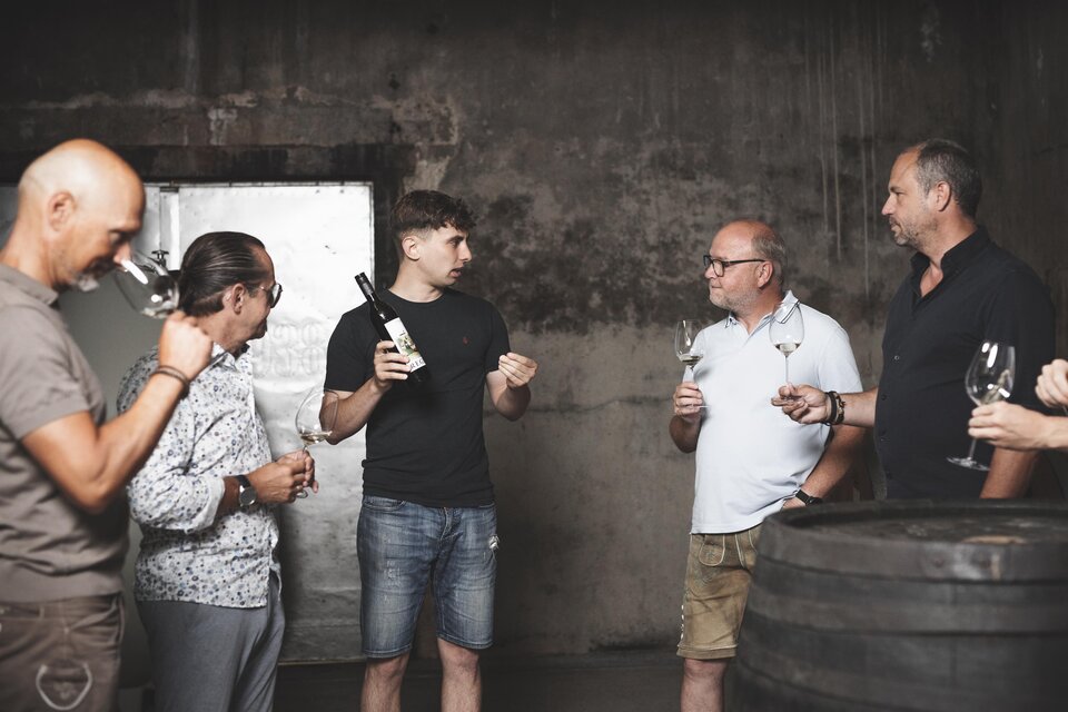 Eine Gruppe von sechs Personen steht in einem Weinkeller. Sie diskutieren und halten Weingläser in der Hand, während einer von ihnen eine Flasche Wein präsentiert. | © Simon Fortmüller