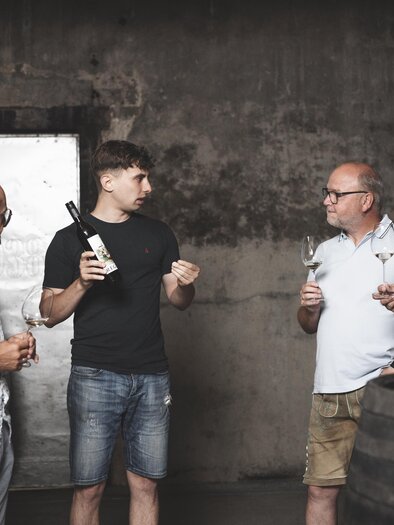 Eine Gruppe von sechs Personen steht in einem Weinkeller. Sie diskutieren und halten Weingläser in der Hand, während einer von ihnen eine Flasche Wein präsentiert. | © Simon Fortmüller