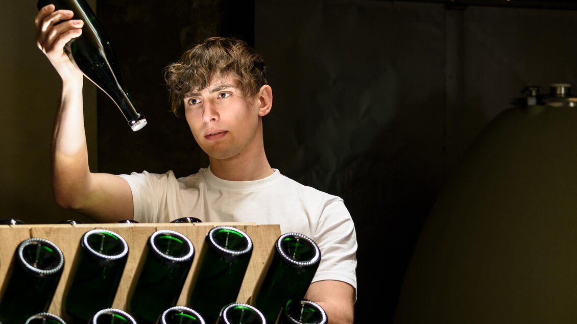 Ein junger Mann prüft eine Flasche in einer Wein- oder Sektkellerei. Im Hintergrund sind mehrere Flaschen in einem Holzgestell zu sehen. | © Mag. Fritz Sutter