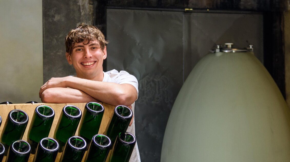 Ein lächelnder junger Mann steht an einer Weinregal mit Flaschen. Im Hintergrund ist ein großer, ovaler Behälter sichtbar. | © Mag. Fritz Sutter