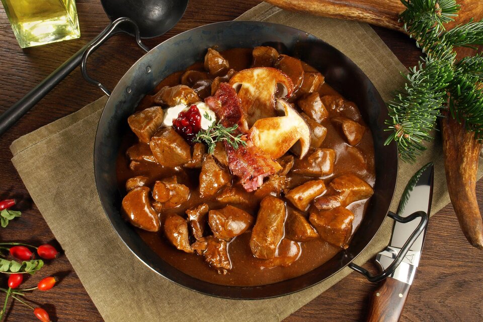 Ein herzhaftes Gericht in einer Pfanne, bestehend aus zartem Fleisch in einer dunklen Sauce. Garniert mit Speck und Kräutern, serviert auf einer rustikalen Tischoberfläche. | © AdobeStock ExQuisine