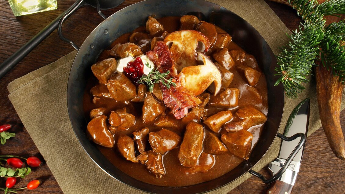 Ein herzhaftes Gericht in einer Pfanne, bestehend aus zartem Fleisch in einer dunklen Sauce. Garniert mit Speck und Kräutern, serviert auf einer rustikalen Tischoberfläche. | © AdobeStock ExQuisine