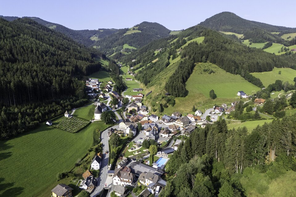 Eine malerische Landschaft mit einem kleinen Dorf umgeben von grünen Hügeln und Wäldern. Der klare Himmel und die sanften Berge schaffen eine friedliche Atmosphäre. | © Kurt Elmleitner