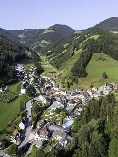 A picturesque landscape with a small village surrounded by green hills and forests. The clear sky and gentle mountains create a peaceful atmosphere. | © Kurt Elmleitner