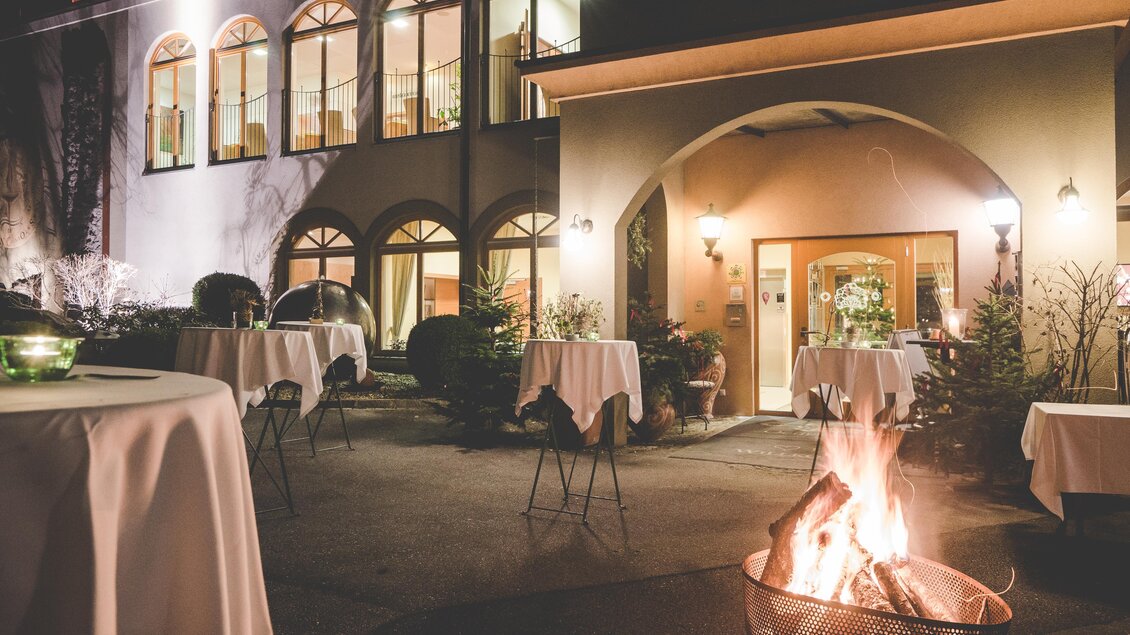 Ein einladender Balkon bei Nacht mit elegant gedeckten Stehtischen. Im Vordergrund brennt ein Lagerfeuer, das für eine warme Atmosphäre sorgt. | © Garten Hotel Ochensberger