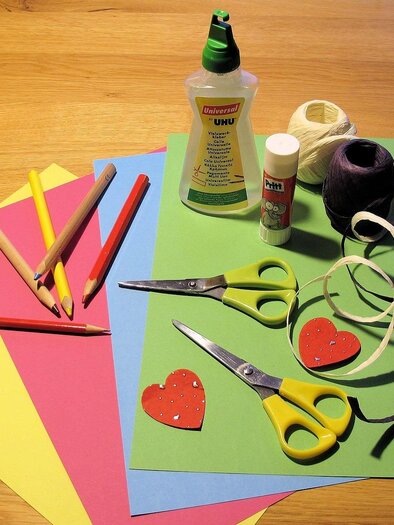 A collection of crafting materials is lying on a wooden table. This includes colored paper, scissors, glue, and colored pencils. | © Pixabay