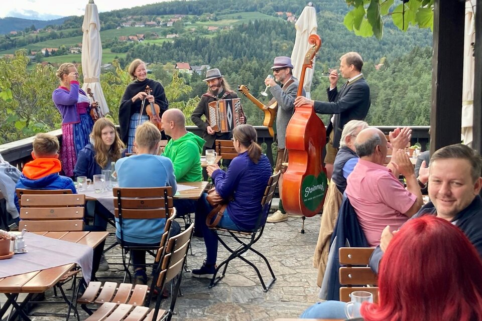 Eine Gruppe von Musikern spielt traditionelle Musik in einem Freiluftrestaurant. Gäste sitzen an Tischen und genießen die Atmosphäre mit Blick auf die Landschaft. | © Weingut Machater