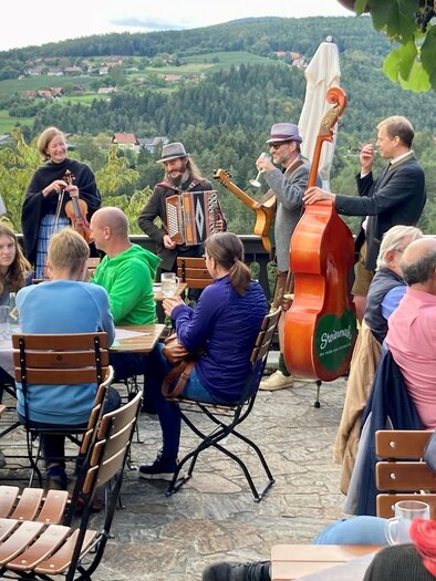 A group of musicians is playing traditional music in an outdoor restaurant. Guests are sitting at tables, enjoying the atmosphere with a view of the landscape. | © Weingut Machater
