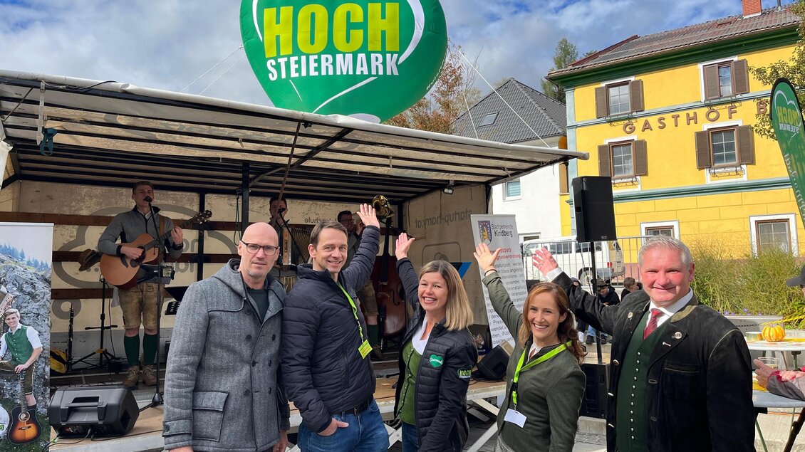Eine fröhliche Gruppe von Menschen steht vor einem Bühnenaufbau mit einem Musiker im Hintergrund. Sie machen Handzeichen und lächeln, während ein großes Schild "HOCH STEIERMARK" über ihnen hängt. | © TV Hochsteiermark