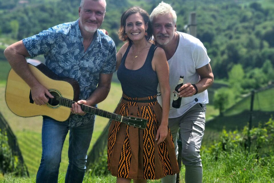 Drei Personen stehen auf einer grünen Wiese mit einer Landschaft im Hintergrund. Ein Mann spielt Gitarre, während die anderen lächeln und sich amüsieren. | © Aperitivo