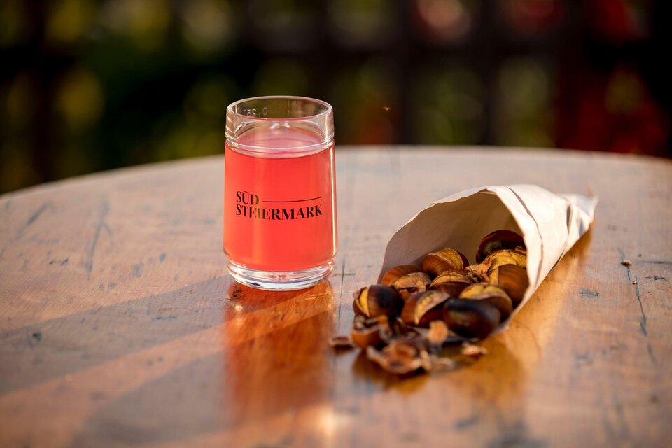 Ein Glas pinkfarbener Drink steht auf einem Holztisch. Neben dem Glas liegt eine Tüte mit gerösteten Kastanien. | © TV Südsteiermark - Tom Lamm