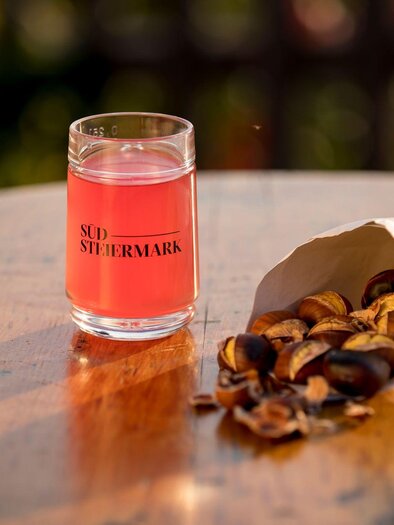 Ein Glas pinkfarbener Drink steht auf einem Holztisch. Neben dem Glas liegt eine Tüte mit gerösteten Kastanien. | © TV Südsteiermark - Tom Lamm