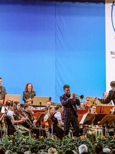 A wind orchestra is playing on stage. The conductor is directing the musicians as the audience listens attentively. | © Blasorchester voestalpine Donawitz