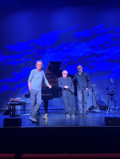 Three musicians are standing on a stage in front of a piano. In the background, a blue lighting design can be seen. | © Köhlmeier
