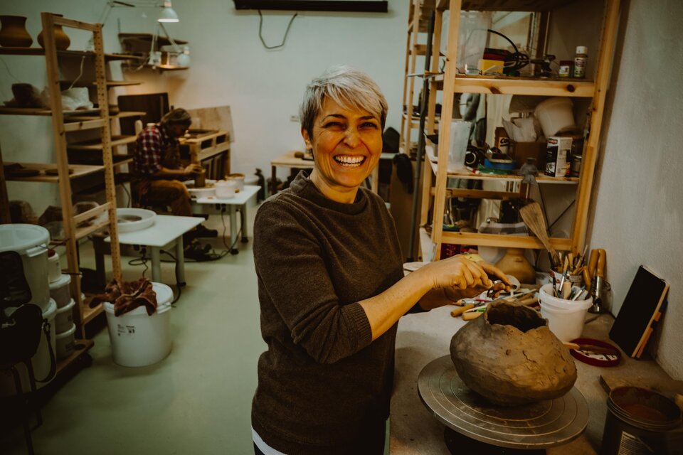 Eine fröhliche Frau arbeitet mit Ton in einem Atelier. Im Hintergrund sind weitere Künstlerin bei der Arbeit zu sehen. | © Karoline Karner