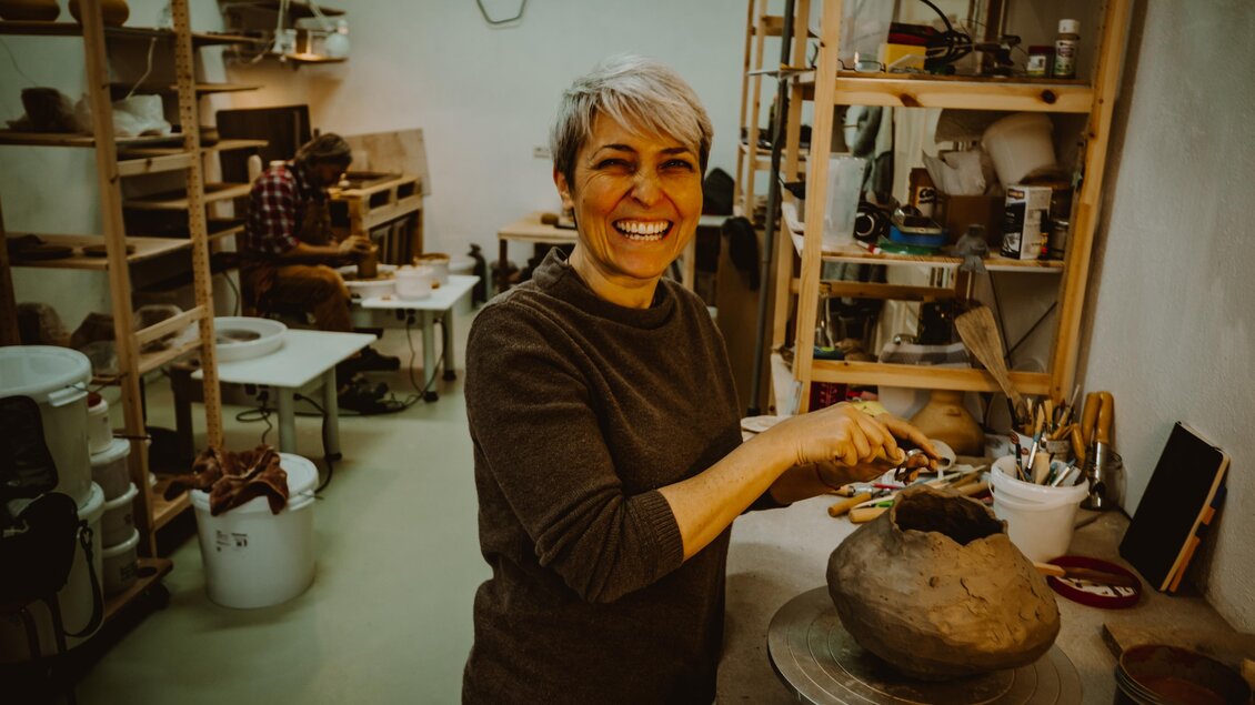 Eine fröhliche Frau arbeitet mit Ton in einem Atelier. Im Hintergrund sind weitere Künstlerin bei der Arbeit zu sehen. | © Karoline Karner