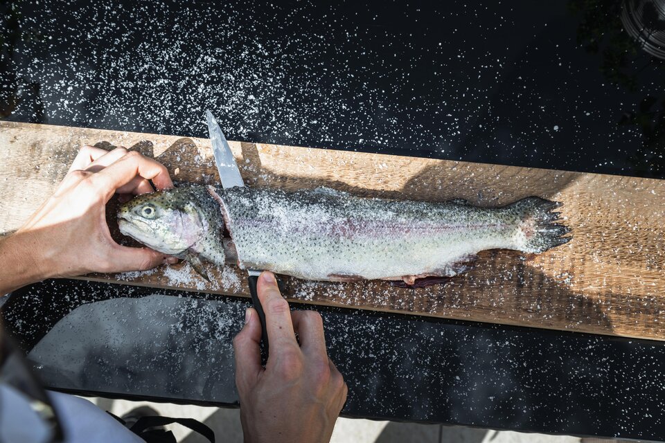 Eine Person schneidet einen frischen Fisch auf einem Holzbrett. Der Fisch liegt auf einer dunklen Oberfläche und ist mit Salz bestreut. | © Vulkanlandhotel Legenstein