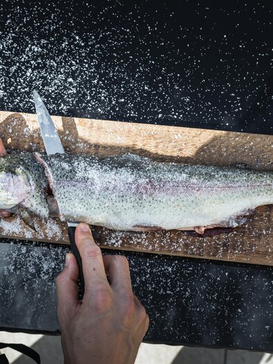 Eine Person schneidet einen frischen Fisch auf einem Holzbrett. Der Fisch liegt auf einer dunklen Oberfläche und ist mit Salz bestreut. | © Vulkanlandhotel Legenstein