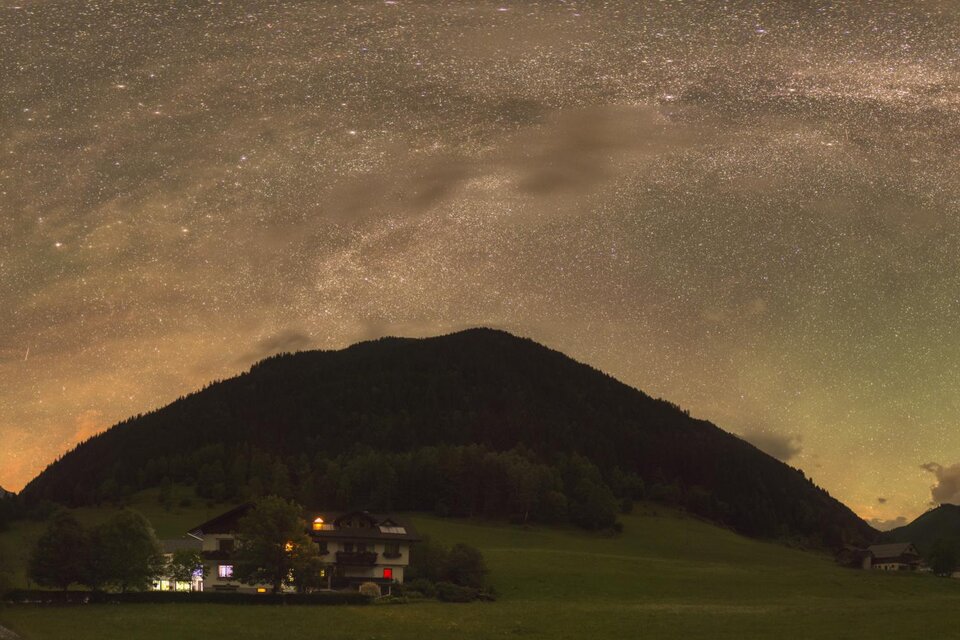 Ein beeindruckender Nachthimmel mit der Milchstraße über majestätischen Bergen. Im Vordergrund sind sanfte Hügel und ein Lichtschein von einem Gebäude zu sehen. | © Zoltán Kolláth