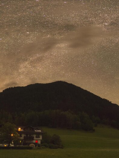 Ein beeindruckender Nachthimmel mit der Milchstraße über majestätischen Bergen. Im Vordergrund sind sanfte Hügel und ein Lichtschein von einem Gebäude zu sehen. | © Zoltán Kolláth
