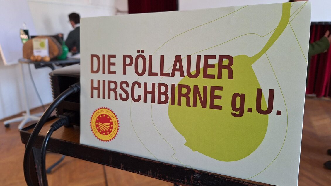 Ein Schild mit der Aufschrift „Die Pöllauer Hirschbirne g.U.“ steht auf einem Tisch. Im Hintergrund sind unscharfe Personen und eine Wand zu sehen. | © Tourismusverband Oststeiermark