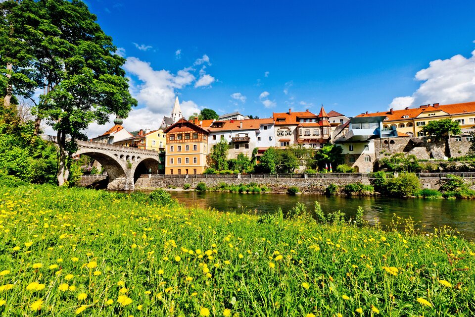 Eine malerische Stadt am Fluss mit bunten Häusern und einer steinernen Brücke. Im Vordergrund blühen gelbe Blumen auf einer Wiese.