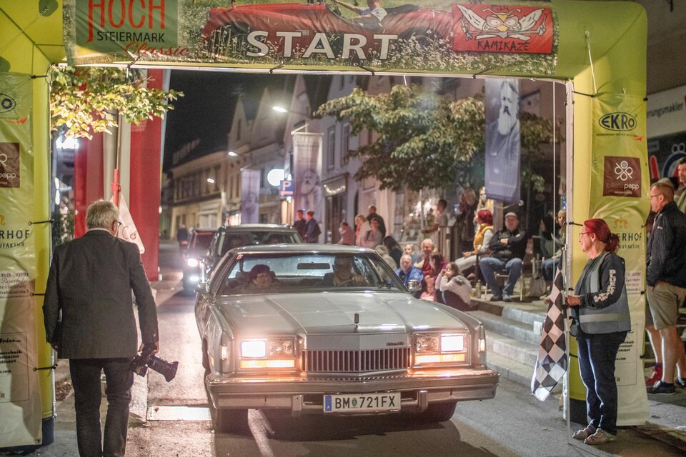Ein klassisches Auto fährt unter einem Startbogen. Um die Straße stehen Zuschauer, die das Ereignis beobachten. | © HOSC Events OG