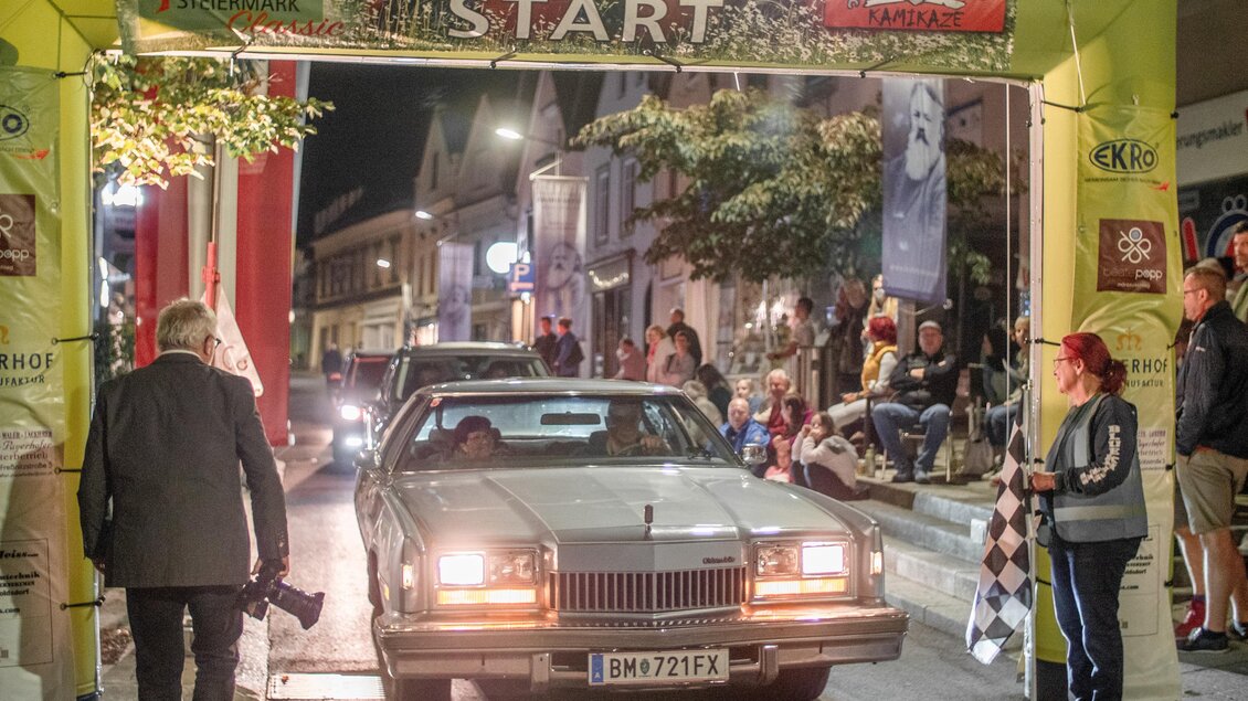 Ein klassisches Auto fährt unter einem Startbogen. Um die Straße stehen Zuschauer, die das Ereignis beobachten. | © HOSC Events OG