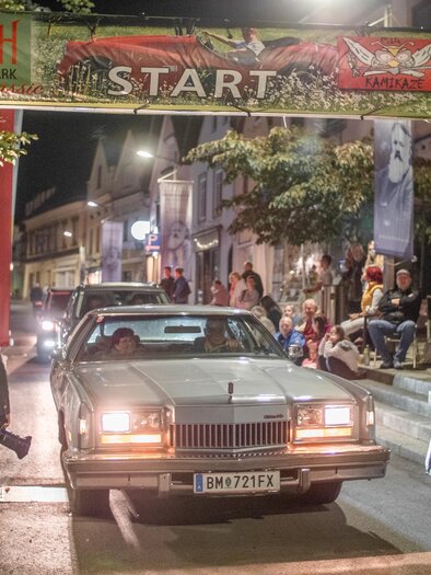 Ein klassisches Auto fährt unter einem Startbogen. Um die Straße stehen Zuschauer, die das Ereignis beobachten. | © HOSC Events OG