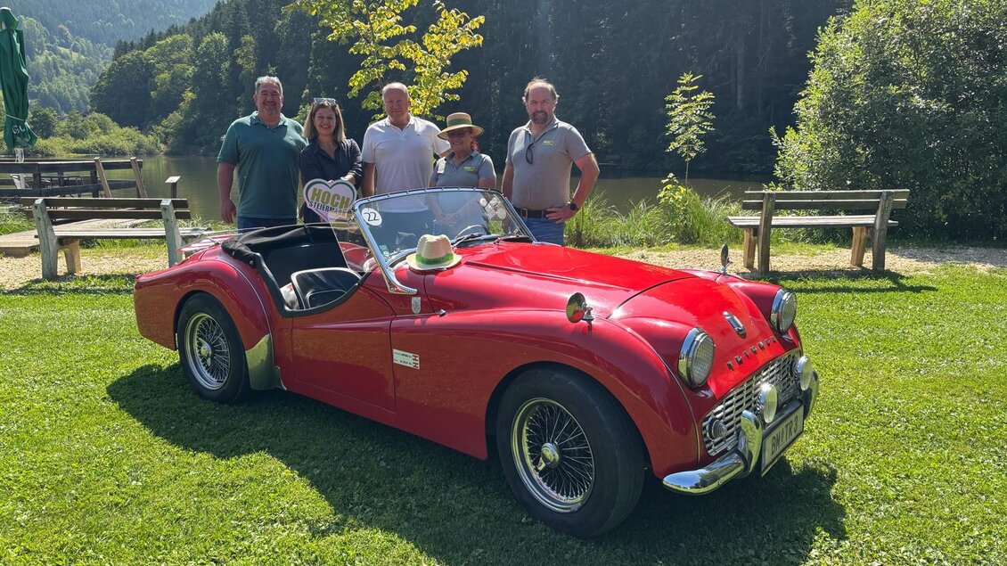Eine Gruppe von fünf Personen steht neben einem roten Sportwagen auf einer grünen Wiese. Im Hintergrund sind Bäume und ein ruhiger See zu sehen.