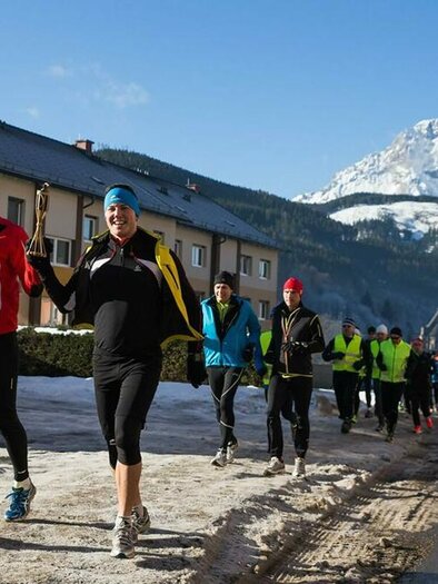 Eine Gruppe von Läufern joggt an einer verschneiten Straße entlang. Im Hintergrund sind Berge und blauer Himmel zu sehen. | © Walter Eisner