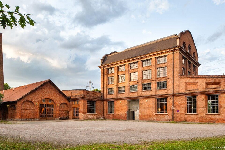 Ein historisches Gebäude aus roten Ziegeln, das früher als Fabrik diente. Im Hintergrund sind graue Wolken und ein Schornstein zu sehen. | © -markus-kaiser-graz.
