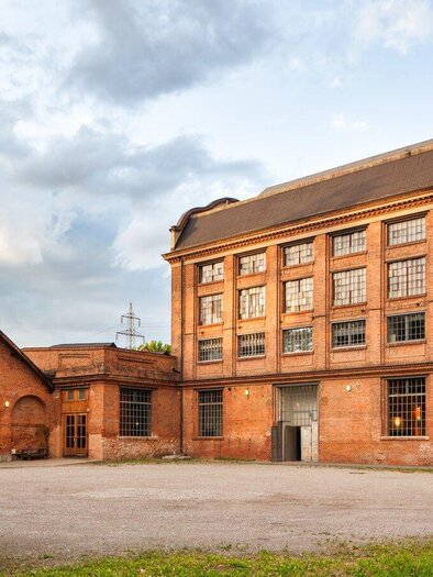 A historic building made of red bricks that used to serve as a factory. In the background, there are gray clouds and a chimney visible. | © -markus-kaiser-graz.