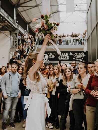 Eine Frau in einem eleganten Outfit hält einen Blumenstrauß hoch. Im Hintergrund steht eine große Menschenmenge, die ihr zusieht. | © hochzeitsfestival-wien