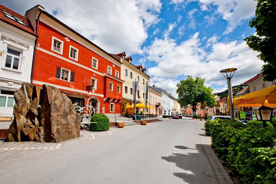 Eine malerische Straße mit bunten Gebäuden und einer großen Steinskulptur. Der Himmel ist klar und die Landschaft wirkt einladend. | © TV Murau