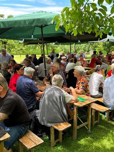 A large group of people is sitting under green sunshades at wooden tables. It looks like a social event outdoors.