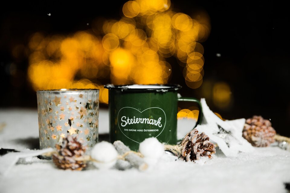 Eine grüne Tasse mit dem Schriftzug "Steiermark" steht neben einem glitzernden Kerzenhalter. Sie sind von Schnee und Tannenzapfen umgeben, mit sanftem Licht im Hintergrund. | © Lupi Spuma