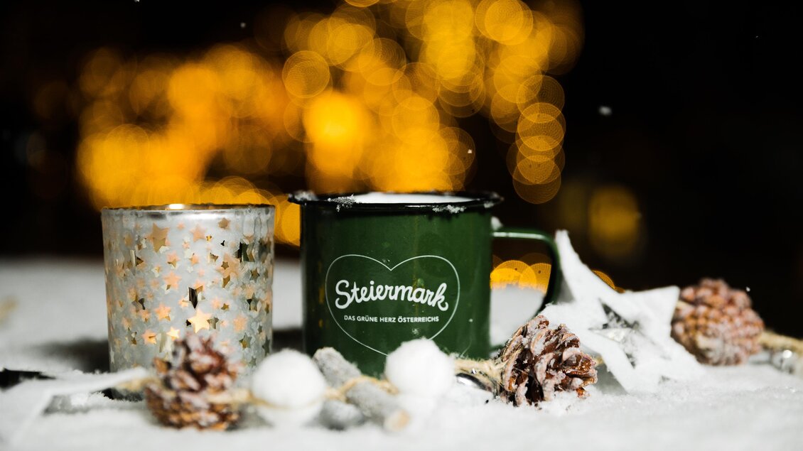 Eine grüne Tasse mit dem Schriftzug "Steiermark" steht neben einem glitzernden Kerzenhalter. Sie sind von Schnee und Tannenzapfen umgeben, mit sanftem Licht im Hintergrund. | © Lupi Spuma