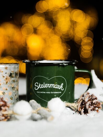 A green mug with the inscription "Styria" stands next to a glittering candle holder. They are surrounded by snow and pine cones, with soft light in the background. | © Lupi Spuma
