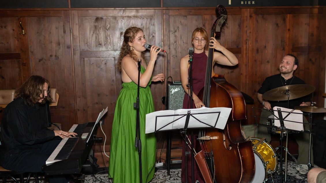 Eine Band spielt in einem gemütlichen Raum. Die Sängerin trägt ein grünes Kleid und wird von weiteren Musikern mit Klavier, Kontrabass und Schlagzeug begleitet. | © Mema Medien Marketing GmbH