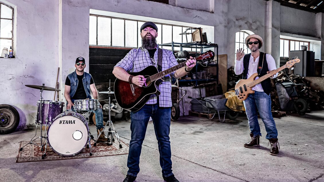 Eine Band spielt in einer Industriehalle. Der Sänger mit Gitarre wird von einem Schlagzeuger und einem Bassisten begleitet. | © Mema Medien Marketing GmbH