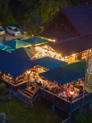 The Hubertus Hut in the evening. The string lights are shining and under the umbrellas, the guests are sitting and enjoying the atmosphere. | © Zechmann